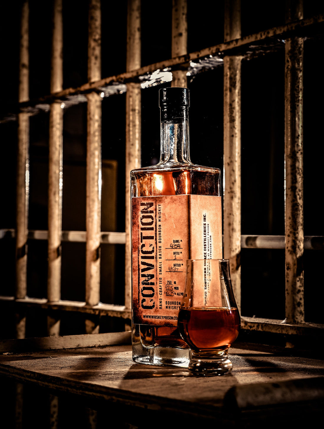 A bottle of Conviction bourbon and a glass in front of old prison bars